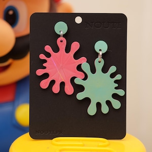 Peut inclure: Boucles d'oreilles avec un motif d'éclaboussure rose et turquoise, fixées à des clous d'oreilles circulaires turquoise. Elles sont présentées sur une carte noire avec le mot "NOUTIER" imprimé.