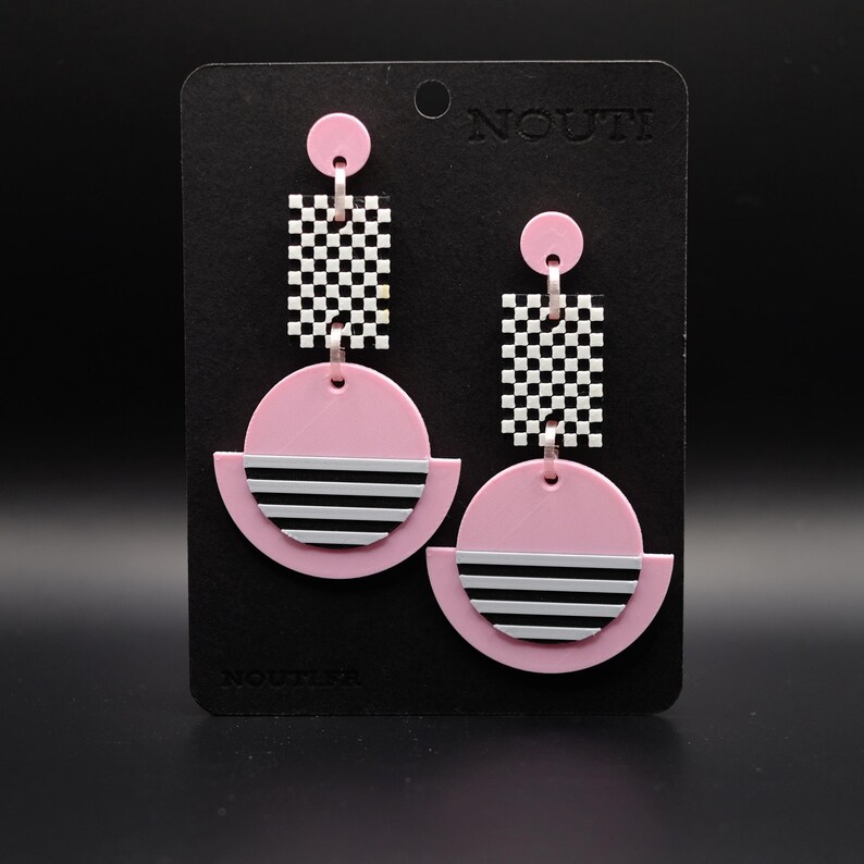 May include: Pink and black geometric earrings with a checkerboard pattern and stripes. The earrings are on a black card with the word "Noutti" printed on it.