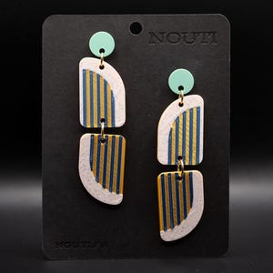 Pode incluir: Um par de brincos geométricos com um design moderno. Os brincos apresentam um esquema de cores bege claro, dourado e azul marinho. Cada brinco tem um pino verde menta, uma seção superior curva e uma seção inferior retangular. A palavra "NOUTI" é impressa no cartão preto.