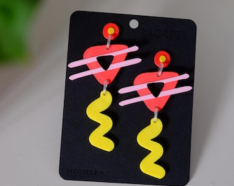 May include: A pair of colorful geometric earrings with pink, red, and yellow accents. The earrings feature a triangle shape with a smaller triangle inside, and a wavy, zig-zag shape hanging below. The earrings are displayed on a black card with the word "NOUITER" printed on it.