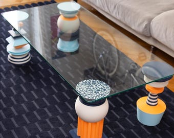 May include: A modern glass-top coffee table with three decorative, colorful pedestal legs. The legs feature geometric shapes and vibrant colors, including orange, blue, and white. The table sits on a dark blue patterned rug.