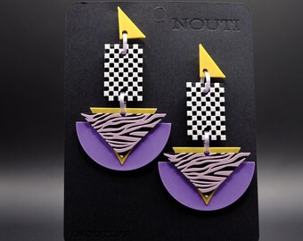 May include: A pair of geometric earrings with a checkerboard, zebra print, and purple and yellow accents. The earrings are displayed on a black card with the word "NOUITI" printed on it.