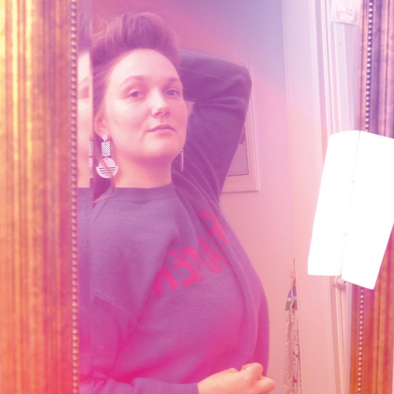 May include: A woman with brown hair is reflected in a mirror. She is wearing a dark blue sweatshirt with a red graphic and large white geometric earrings.