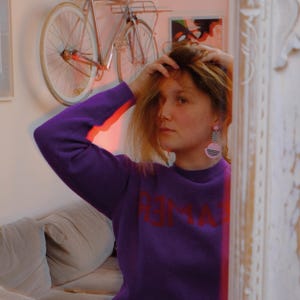 May include: A woman with long blonde hair wearing a purple sweater with the word "REMARK" printed in red on the front. She is wearing large pink and black geometric earrings.