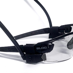 May include: Black sports sunglasses with clear lenses. The sunglasses have a wraparound design and a strap that goes around the head. The brand name "BLOC" is visible on the frame. The lenses are clear and the frame is black.