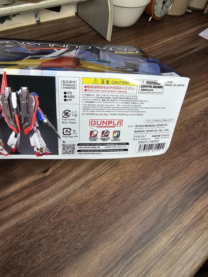 May include: A box for a RG 1/144 ZETA GUNDAM plastic model kit. The box features a detailed image of the assembled model, showcasing its red, white, blue, and gray color scheme. The box also includes Japanese text and warnings.