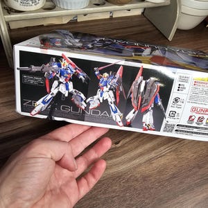 May include: A box featuring a Gundam model kit. The box art shows three Gundam figures in dynamic poses, with a color scheme of white, blue, red, and gray. The text "ZE GUNDA" is visible.