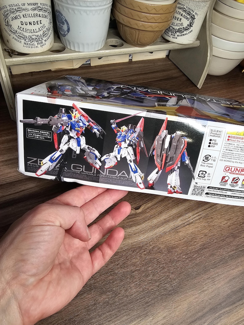 May include: A box featuring a Gundam model kit. The box art shows three Gundam figures in dynamic poses, with a color scheme of white, blue, red, and gray. The text "ZE GUNDA" is visible.