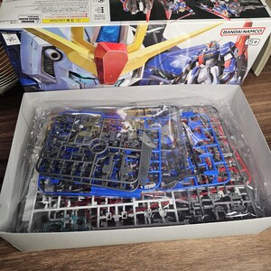 May include: A Bandai Namco Gundam model kit box, featuring a detailed illustration of a blue, red, and yellow mecha. The box is open, revealing plastic model parts in various colors. The kit is marked for ages 15+.