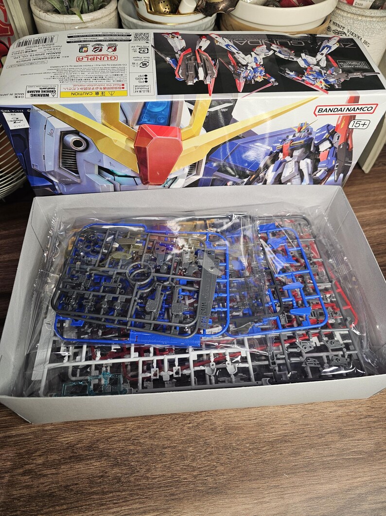 May include: A Bandai Namco Gundam model kit box, featuring a detailed illustration of a blue, red, and yellow mecha. The box is open, revealing plastic model parts in various colors. The kit is marked for ages 15+.