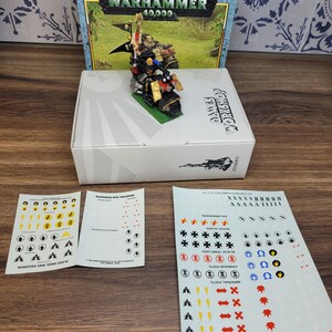 May include: A miniature plastic model of a Space Marine riding a motorcycle, with a red and yellow color scheme. The model is part of the Warhammer 40,000 game.