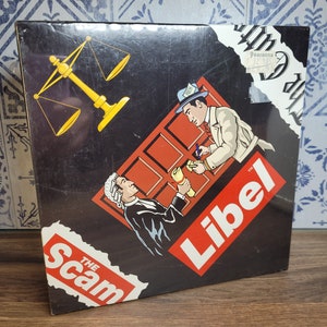 Pode incluir: Uma caixa de jogo de tabuleiro vintage com fundo preto e ilustrações. O jogo é intitulado "The Scam Libel" com imagens de um juiz, balanças da justiça e um detetive. A caixa tem uma aparência desgastada.