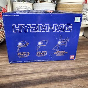 Może przedstawiać: Niebieskie pudełko z napisem "HY2M-MG" i ilustracjami głów robotów. Pudełko zawiera również tekst "MS-06-1A ZAKU-II", "MS-05B ZAKU-I" i "MS-14S GELGOOG". Widoczne jest logo Bandai.