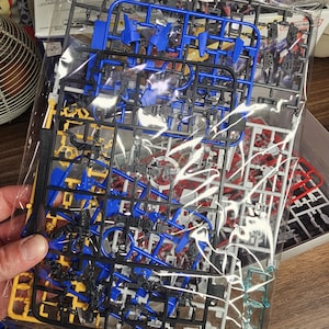 May include: A clear plastic bag containing unassembled model kit parts. The parts are various colors, including blue, yellow, black, red, and white. The parts are attached to plastic frames, ready for assembly. The kit appears to be for a robot or vehicle model.