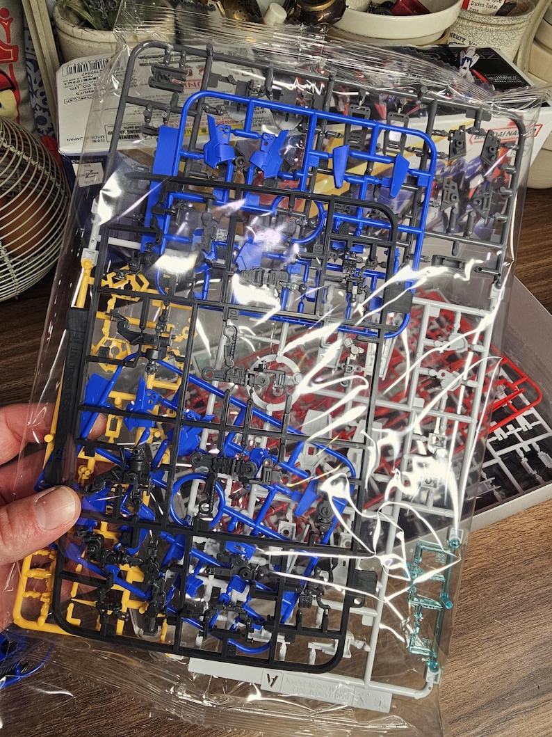 May include: A clear plastic bag containing unassembled model kit parts. The parts are various colors, including blue, yellow, black, red, and white. The parts are attached to plastic frames, ready for assembly. The kit appears to be for a robot or vehicle model.