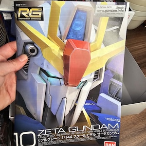 May include: A box for a Zeta Gundam model kit. The box features a detailed illustration of the robot's head in shades of white, blue, gold, and red. Text includes "Zeta Gundam" and "1/144 scale model". The box is held by a person.