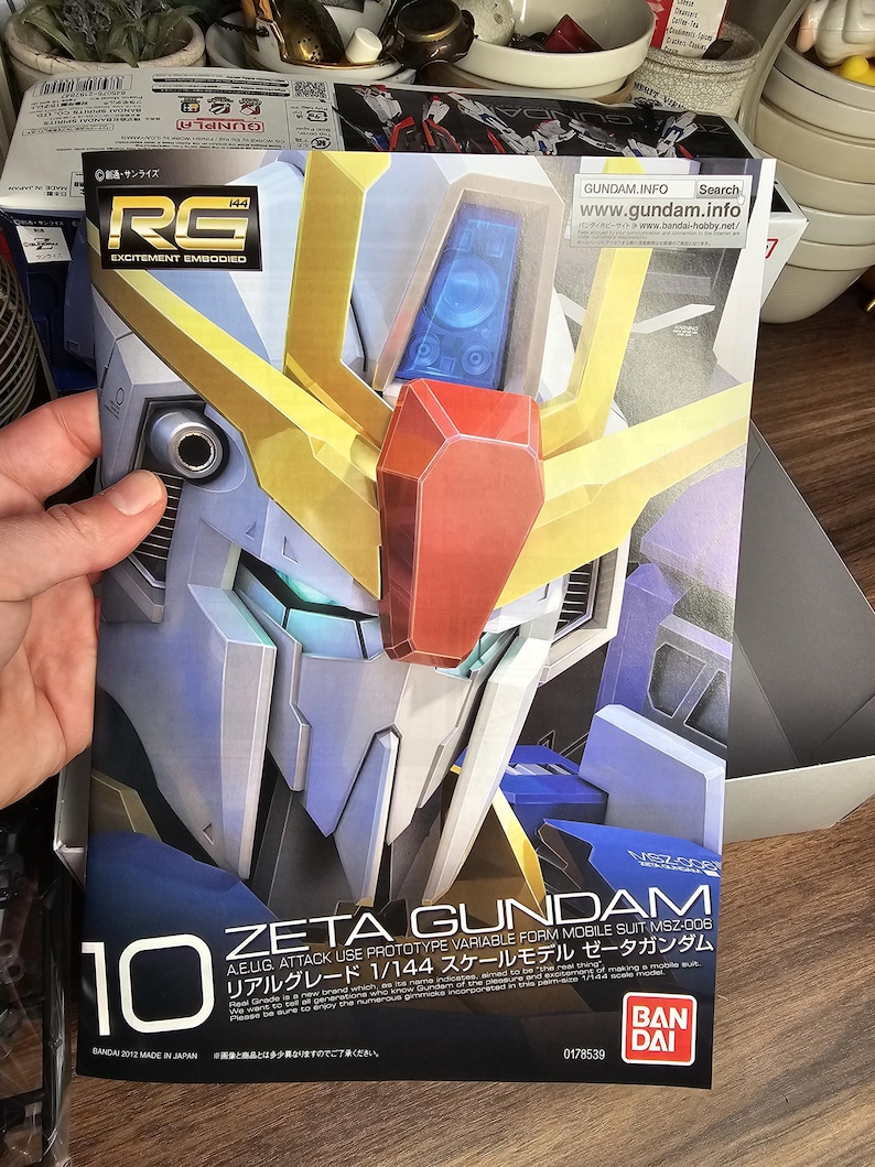 May include: A box for a Zeta Gundam model kit. The box features a detailed illustration of the robot's head in shades of white, blue, gold, and red. Text includes "Zeta Gundam" and "1/144 scale model". The box is held by a person.