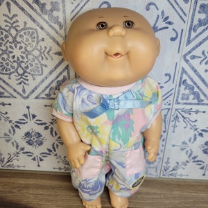 Peut inclure: Une poupée Cabbage Patch Kid avec une tête chauve, des yeux bruns et une tenue à fleurs rose et bleue. La poupée porte un ruban bleu autour du cou.