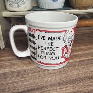 May include: White ceramic mug with a black and red design. The mug features the text "I'VE MADE THE PERFECT THING FOR YOU" with a cartoon character illustration. The mug has a large handle and is set against a wooden surface.