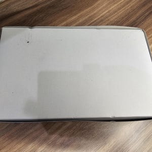 May include: A rectangular, light gray cardboard box with a slightly textured surface. The box is positioned on a wooden surface with a visible wood grain pattern. The box is closed and appears empty.