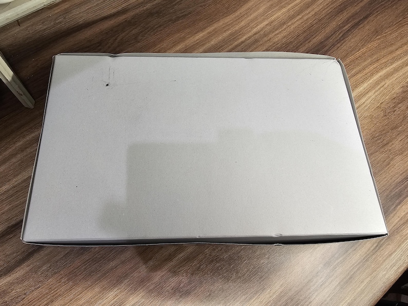 May include: A rectangular, light gray cardboard box with a slightly textured surface. The box is positioned on a wooden surface with a visible wood grain pattern. The box is closed and appears empty.