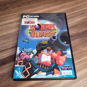 Puede incluir: Caja de juego para PC CD-ROM de Worms Blast. La portada presenta un gusano de dibujos animados con gafas sosteniendo una pistola grande, sobre un fondo de isla tropical. El título del juego se muestra de forma destacada con las palabras "Worms Blast".