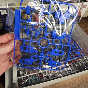 May include: A blue plastic model kit sprue, held in a clear plastic bag. The sprue contains various parts for a model, with a focus on mechanical components. The kit is partially assembled, with other parts visible in the box.