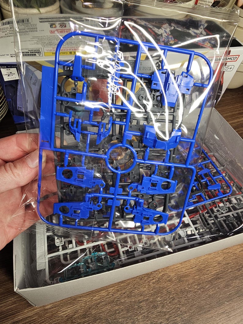 May include: A blue plastic model kit sprue, held in a clear plastic bag. The sprue contains various parts for a model, with a focus on mechanical components. The kit is partially assembled, with other parts visible in the box.
