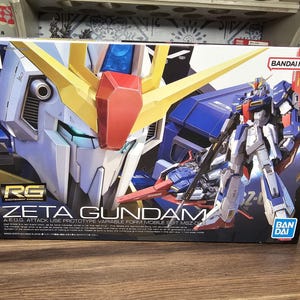 May include: A box featuring the Zeta Gundam model kit. The box art showcases a detailed illustration of the robot in blue, white, yellow, and red. The text "ZETA GUNDAM" is prominently displayed, along with the "BANDAI NAMCO" logo.