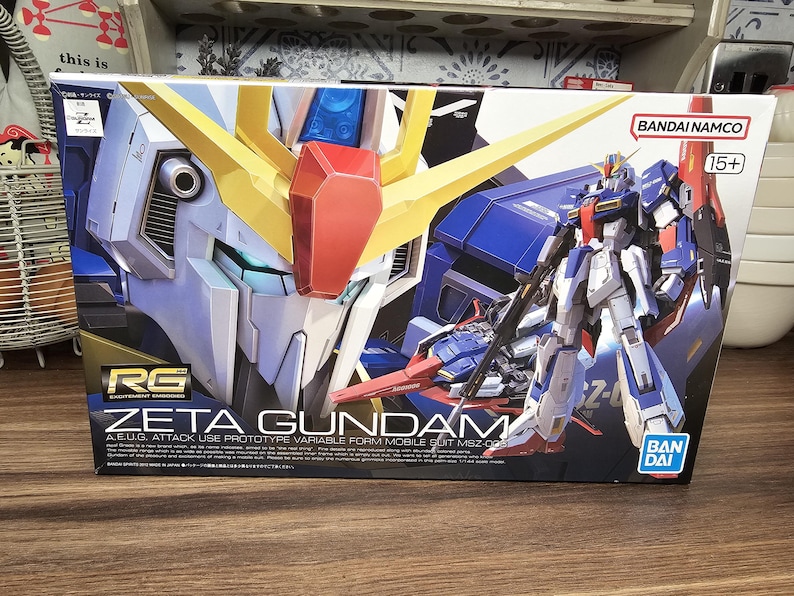 May include: A box featuring the Zeta Gundam model kit. The box art showcases a detailed illustration of the robot in blue, white, yellow, and red. The text "ZETA GUNDAM" is prominently displayed, along with the "BANDAI NAMCO" logo.