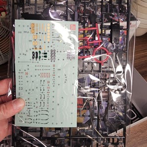 May include: A plastic model kit, featuring black and red components, sealed in a clear plastic bag. The kit includes a sheet of decals with various markings and details. The parts are arranged on black plastic frames, ready for assembly.