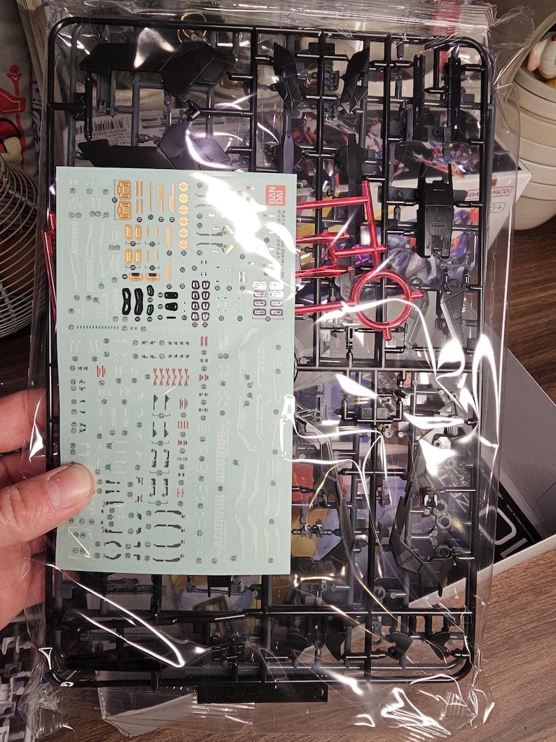 May include: A plastic model kit, featuring black and red components, sealed in a clear plastic bag. The kit includes a sheet of decals with various markings and details. The parts are arranged on black plastic frames, ready for assembly.