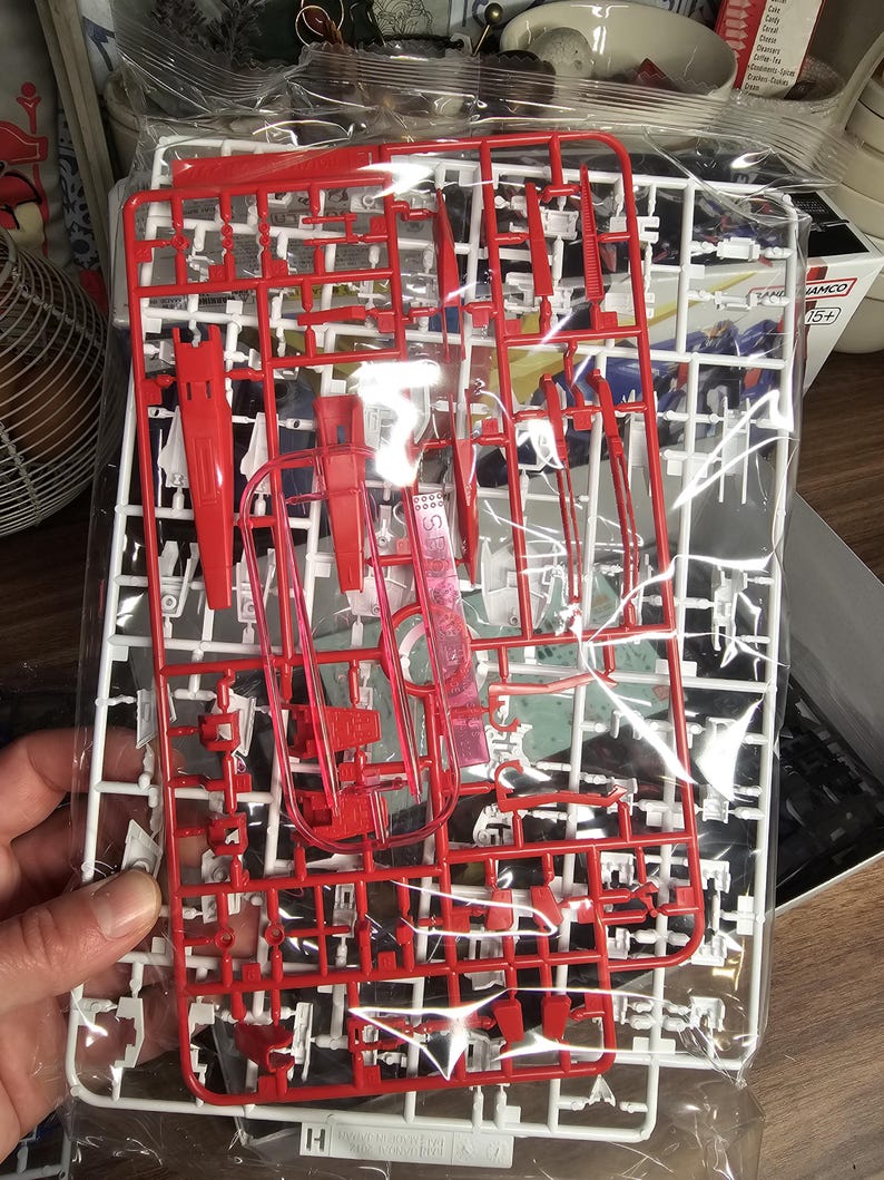 May include: A clear plastic bag containing red and white plastic model kit parts. The parts are attached to a white frame, with various shapes and sizes. The kit appears to be for a robot or vehicle model.