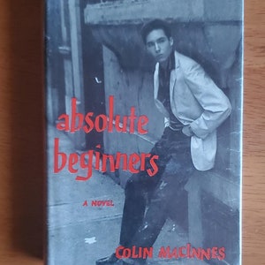 May include: A book cover with a black and white photo of a young man in a white jacket. The title of the book is "absolute beginners" in red lettering. The author's name, "COLIN MACINNES", is also in red lettering.