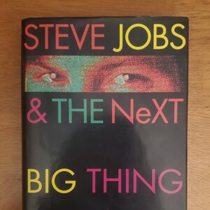 May include: A hardcover book titled "STEVE JOBS & THE NeXT BIG THING" by Randall E. Stross. The cover features a black background with the title in various colors and a close-up of eyes.