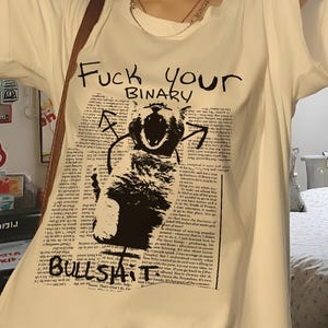 Peut inclure: T-shirt blanc avec une impression graphique et le texte "Fuck your BINARY BULLSHIT". Le t-shirt a des manches courtes et un col rond. Le graphisme est une illustration en noir et blanc.