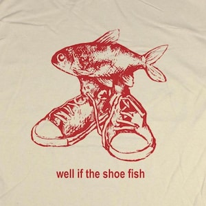 May include: Cream-colored t-shirt featuring a red graphic of a fish atop a pair of sneakers. The text "well if the shoe fish" is printed below the graphic. The shirt has short sleeves and a crew neck.