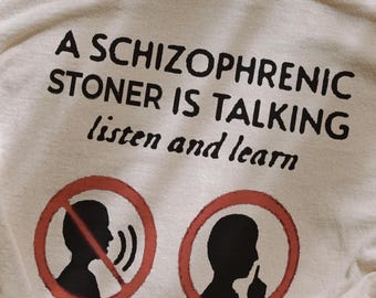 May include: A beige t-shirt with black text that reads "A Schizophrenic Stoner is Talking listen and learn". The shirt has two red circles with black silhouettes of people's heads. One person has a sound wave coming out of their mouth and the other person has their finger over their mouth.