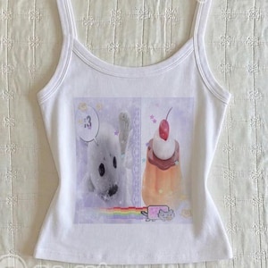 May include: A white sleeveless tank top featuring a central graphic print. The image displays a white seal, a dessert topped with a cherry, a small cat icon, and a rainbow. The text :3 is visible in a speech bubble above the seal.