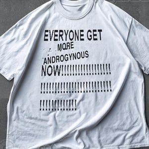 Könnte beinhalten: Weißes T-Shirt mit dem Text "EVERYONE GET MORE ANDROGYNOUS NOW!!!!" in Schwarz. Das Shirt hat einen Rundhalsausschnitt und kurze Ärmel. Der Text ist mittig auf der Vorderseite des Shirts platziert.