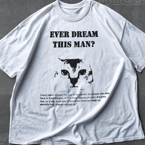 May include: A white short-sleeve t-shirt featuring the black text EVER DREAM THIS MAN? above a graphic of a cat face. Below the cat is the text: Every night, all over the world, hundreds of people see this face in their dreams.