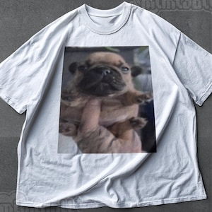 Pode incluir: Camiseta branca com uma imagem impressa de um filhote marrom. O filhote está sendo segurado, mostrando seu rosto e patas. A camiseta tem gola redonda e mangas curtas.