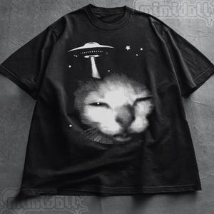 May include: Black t-shirt with a white graphic of a cat's face and a UFO. Stars are also included in the design. The short-sleeved tee has a crew neck. The design is a striking contrast against the dark fabric.