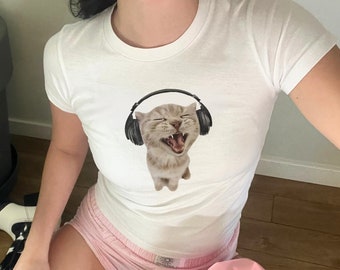 May include: A white t-shirt with a graphic of a cat wearing headphones. The cat is singing and has its mouth open. The person wearing the shirt is sitting on the floor with their legs crossed. They are wearing pink shorts and white boots.