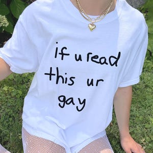 May include: White t-shirt with the handwritten text "if u read this ur gay" in black. The shirt is paired with white shorts and fishnet stockings. A gold necklace with a heart pendant is visible.