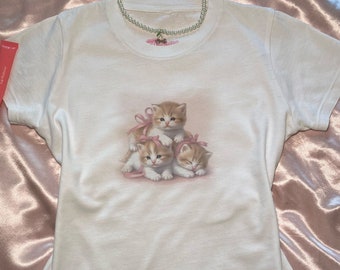 Puede incluir: Camiseta blanca con un gráfico de tres gatitos con lazos rosas. Los gatitos están apilados uno encima del otro.