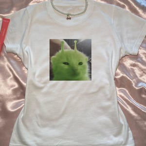 以下が含まれることがあります： 緑色の宇宙人猫のグラフィックプリントが施された白いTシャツ。猫はアンテナがあり、真剣な表情をしています。