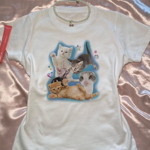 Peut inclure: T-shirt blanche avec un imprim&eacute; graphique de cinq chatons dans une pose ludique. Les chatons sont entour&eacute;s de c&oelig;urs, d'&eacute;toiles et d'empreintes de pattes color&eacute;s.