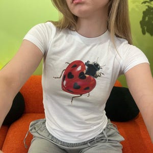 Puede incluir: Camiseta blanca con un gráfico de una mariquita con manchas negras y un cuerpo rojo en forma de corazón.