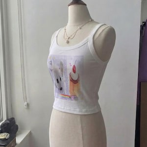 May include: A white sleeveless crop top featuring a graphic print of a white creature, a cupcake with a cherry on top, and a small cat on a rainbow. The top has a scoop neckline and is displayed on a mannequin wearing a delicate pearl necklace.